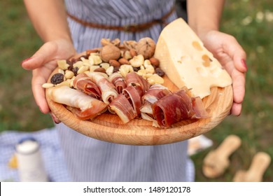 Young Woman Holds A Wooden Dish With Delicacies. Snacks For A Summer Picnic Jamon, Cheese, Dry Fruits, Jerky And Nuts.