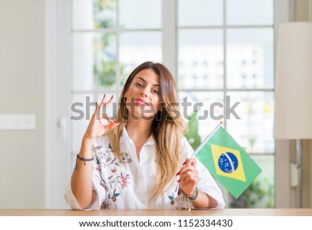 Young woman at home holding flag of Brazil doing ok sign with fingers, excellent symbol