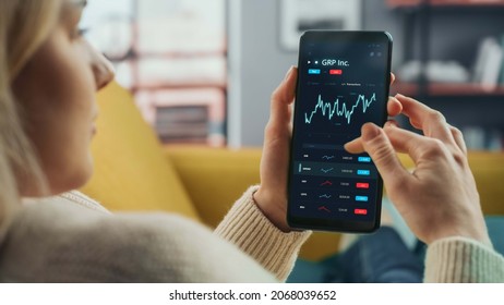 Young Woman At Home Lying On A Couch And Using Smartphone With Stock Market App To Check Share Prices. Graph Is Going Up Making A Great Profit. In The Background Cozy Homely Atmosphere.