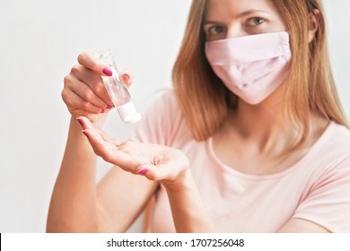 Young Woman With Home Made Pink Cotton Face Mouth Virus Mask Dropping Anti Bacterial Alcohol Rubbing Gel On Hand, Detail To White Bottle, Blurred Face In Background. Coronavirus Covid-19 Prevention