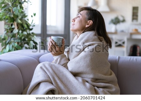 Young woman at home on the sofa in winter wrapped herself in a warm cozy blanket, holding hot cocoa coffee