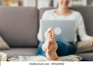 Young Woman At Home Relaxing On The Couch, She Is Using A Laptop, Feet Up On The Foreground