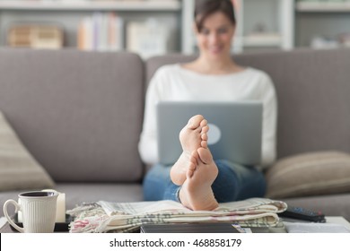 Young Woman At Home Relaxing On The Couch, She Is Using A Laptop, Feet Up On The Foreground