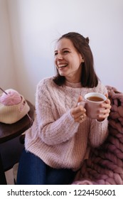 Young Woman At Home Sitting On Chair Relaxing, Drinking Tea. Soft Cozy Photo Of Slim Girl In Warm Sweater With Cup Of Tea In Hands. Girl Sitting On Giant Knitted Plaid. Knitting, Wool Thread Needles