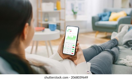 Young Woman At Home Sitting On A Couch And Using Smartphone With Stock Market App To Check Share Prices. Graph Is Going Up Making A Great Profit. In The Background Cozy Homely Atmosphere.