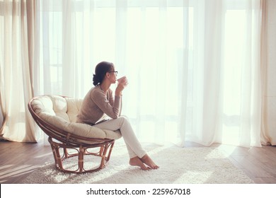 Young Woman At Home Sitting On Modern Chair In Front Of Window Relaxing In Her Living Room And Drinking Coffee Or Tea