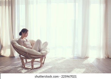 Young Woman At Home Sitting On Modern Chair In Front Of Window Relaxing In Her Living Room Using Tablet Pc