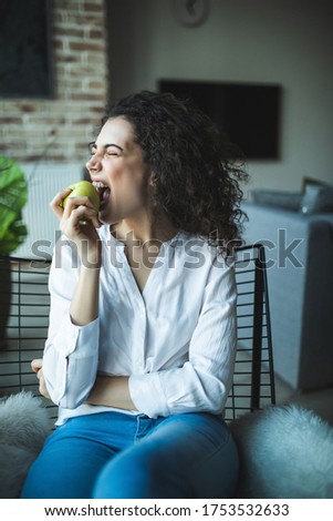Young woman at home sitting on modern chair eat apple in front of window relaxing at home