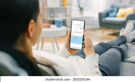 Young Woman at Home Uses Smartphone for Checking Weather Predictions and Coming Day Plans in Daily Schedule Calendar. She's Sitting On a Couch in Her Cozy Living Room. Over the Shoulder Shot - Shutterstock ID 1833205144