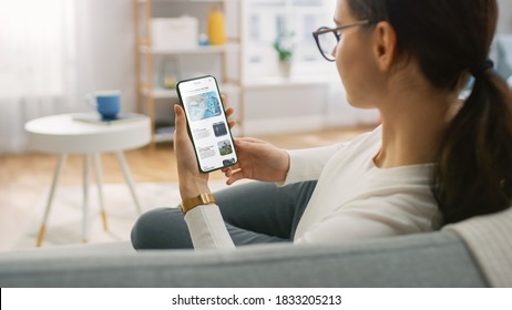 Young Woman at Home Uses Smartphone for Scrolling and Reading News about Technological Breakthroughs. She's Sitting On a Couch in Her Cozy Living Room. Over the Shoulder Shot - Powered by Shutterstock