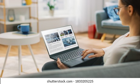 Young Woman at Home Is Using Laptop Computer for Scrolling and Reading News about Technological Breakthroughs. She's Sitting On a Couch in His Cozy Living Room. Over the Shoulder Shot
