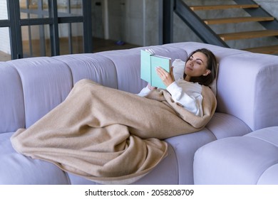 A young woman at home in a white hoodie on the couch wrapped herself in a warm blanket, reads a book, a novel, winter evening