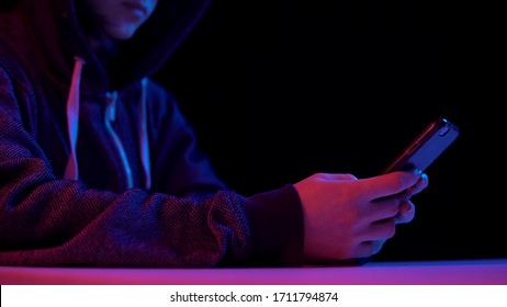 Young Woman In A Hood With A Smartphone. Hacker Makes A Hack Through The Phone. Blue And Red Light Falls On A Woman On A Black Background.