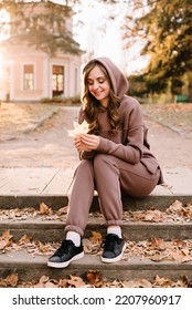 Young Woman In Hoodie Sitting On The Steps In An Autumn Park. Sunny Weather. Fall Season.