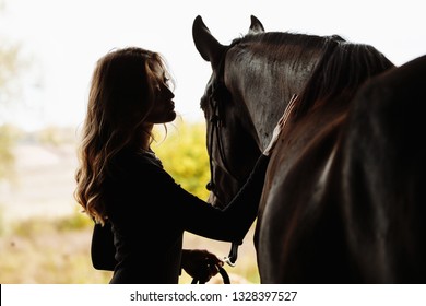 young woman with horse in stable silhouetted with the grass and farm behind her