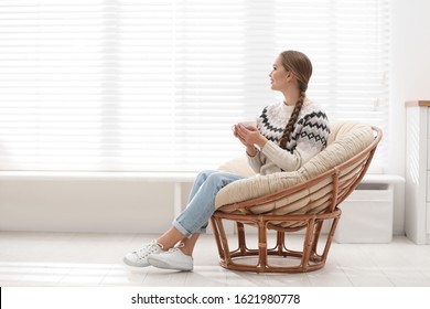Young woman with hot drink in papasan chair near window at home