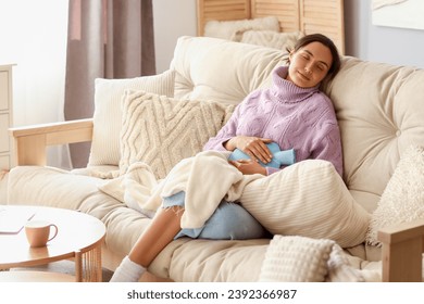 Young woman with hot water bottle sitting on sofa at home - Powered by Shutterstock