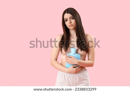 Young woman with hot water bottle having menstrual cramps on pink background