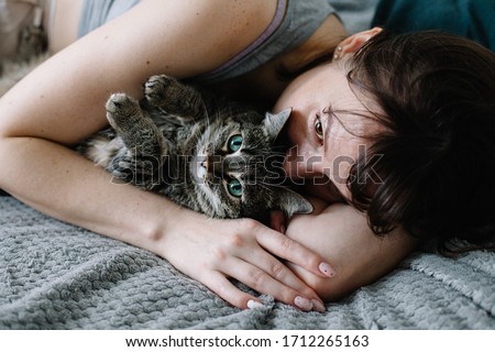 Young woman hug her cat on the bed at home. woman and cat lie on a gray bedspread against a gray wall. the woman has a brunette. cat in the arms of the mistress.sad woman look. the cat lies on its bad