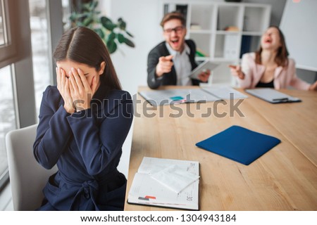 Young woman hurt by rude colleagues. She sit at table near windown and close her face with hands. People scream and laughing on her. They behave bad.