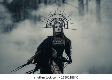 Young woman in image of witch stands in black dress and crown on her head in dark and foggy forest.