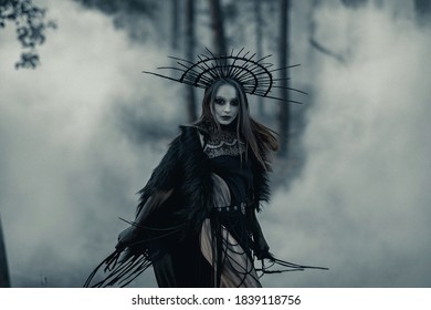 Young woman in image of witch walks in black dress and crown on her head across foggy forest.