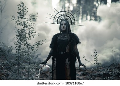 Young woman in image of witch walks in black dress and crown on her head with human skull in her hand through smoke in forest.