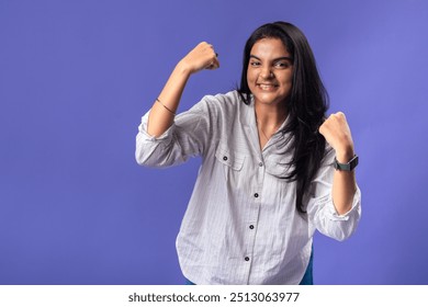 A young woman of Indian descent, wearing a white striped shirt and black smartwatch, stands against a solid purple background with her arms raised in a victory pose celebrating success - Powered by Shutterstock