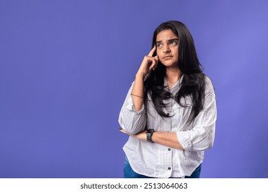 A young woman of Indian descent, wearing a white striped shirt and black smartwatch, poses thoughtfully with her finger on her temple, thought against a solid purple background - Powered by Shutterstock