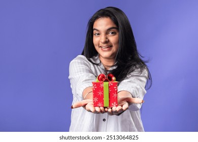 A young woman of Indian descent, wearing a white striped shirt and black smartwatch, stands against a solid purple background, smiling brightly while holding out a festive red gift box - Powered by Shutterstock