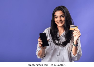 A young woman of Indian descent, wearing a white striped shirt and black smartwatch, stands against a solid purple background, smiling while holding a smartphone in one hand and a credit card - Powered by Shutterstock