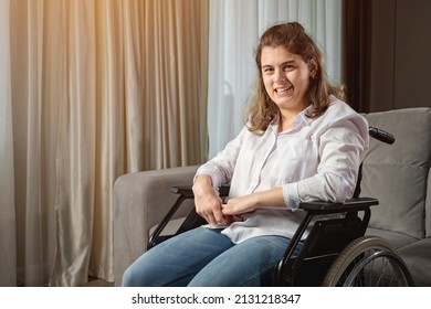 Young woman with infantile cerebral paralysis in white shirt smiles sitting in wheelchair near stylish sofa in living room at home, sunlight.