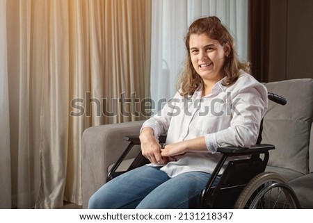 Young woman with infantile cerebral paralysis in white shirt smiles sitting in wheelchair near stylish sofa in living room at home, sunlight.