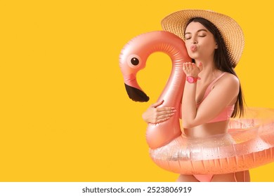 Young woman with inflatable ring in shape of flamingo blowing kiss on yellow background - Powered by Shutterstock