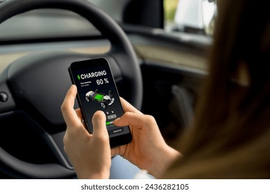 Young woman inside the vehicle, checking EV vehicle's application on battery recharging electricity status display on smartphone screen in modern EV car on her road trip journey. Exalt - Powered by Shutterstock