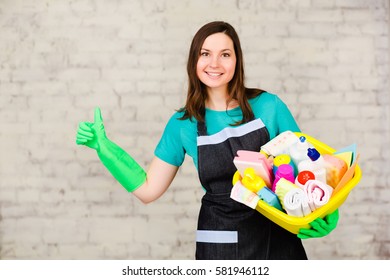 Young woman janitor holding bucket with many colorful detergent for cleaning office. Worker cleaning service. On the soft-focus background a white brick wall