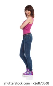 Young Woman In Jeans, Pink Tank Top And Sneakers Is Standing, Holding Arms Crossed, Looking At Camera And Smiling. Side View. Full Length Studio Shot Isolated On White.