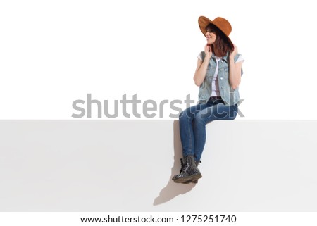 Young woman in jeans vest, black boots and brown suede hat is sitting on a top, looking away and smiling. Full length studio shot isolated on white.