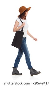 Young Woman In Jeans Vest, Boots And Brown Suede Hat Is Walking, Holding Black Bag On Her Shoulder And Looking Away. Side View. Full Length Studio Shot Isolated On White.