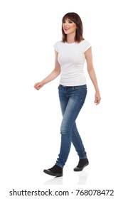 Young woman in jeans and white t-shirt is walking, looking away and talking. Full length studio shot isolated on white.