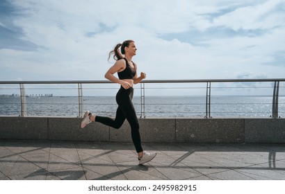 young woman jogging along a waterfront path, dressed in black sportswear. The sky is partly cloudy, and she appears focused and determined, enjoying her outdoor run - Powered by Shutterstock