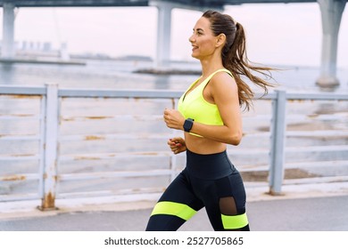 Young Woman Jogging Along a Waterfront Promenade in Bright Workout Clothes During the Day Under a Bridge - Powered by Shutterstock