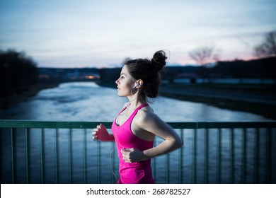 Young Woman Jogging At Night In The City On The Bridge Cross The River. Girl Running Outdoors In A City Park. Motion Blurred Image. Color Toned Image. 