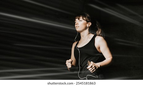 Young woman jogging outdoors, wearing headphones. Focused female runner in motion, enjoying exercise. Athletic woman, active lifestyle, fitness enthusiast. Workout, health concept with copy space. - Powered by Shutterstock