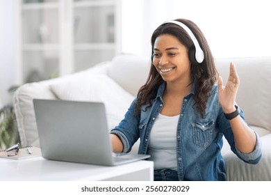 A young woman is joyfully engaging in a video call from her cozy living room, wearing headphones and a casual outfit. Natural light fills the space, enhancing the cheerful atmosphere. - Powered by Shutterstock