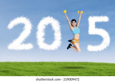 Young woman jumping on meadow while holding two dumbbells and forming number 2015