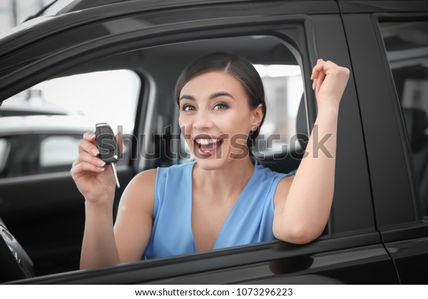 Young woman with key sitting in driver\'s seat of\
new car at salon