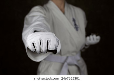 Young woman in kimono and white belt throws punch in protective gloves in Kyokushin karate to camera, focus on fist with blurred martial arts athlete on background. Inscription in Japanese - Powered by Shutterstock