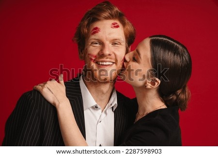 Young woman kissing cheerful man with red lipstick marks on his face isolated over red studio wall
