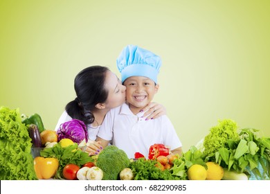 Young Woman Kissing Her Son While Preparing Fresh Vegetables On The Table, Shot With Green Screen Background